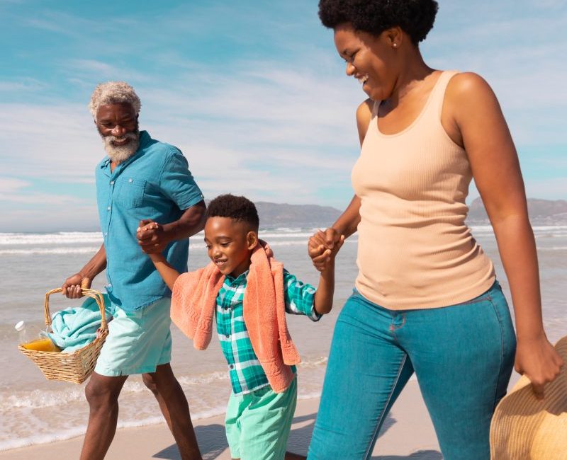 Happy african american grandparents holding boy's hands and walking against sea and sky in summer. nature, basket, hat, unaltered, family, love, togetherness, childhood, enjoyment, holiday concept. | Holidays to california for couples & families